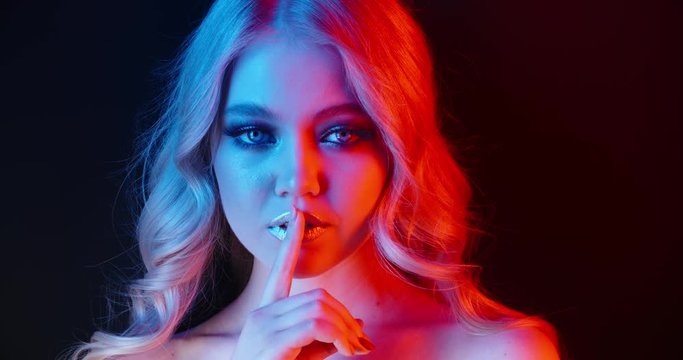 Beautiful caucasian girl wearing glowing makeup in neon light sensually looking at camera and pressing finger against her lips - nightlife, cybcerpunk concept 4k footage