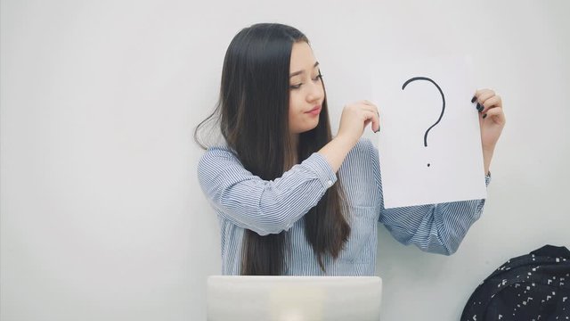 Lovely young asian lady sitting, with laptop, raising a sheet of paper with question mark written on it, nodding her head, understanding nothing. Cheerless face expression.