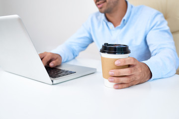 Young entrepreneur drinking cup of take away coffee when working on laptop in office