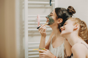 Side view portrait of two girlfriends doing facial masks in the bathroom smiling.