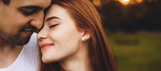 Close up portrait of a beautiful young couple sitting face to face with eyes closed smiling before kissing while man is embracing her from back against sunset.