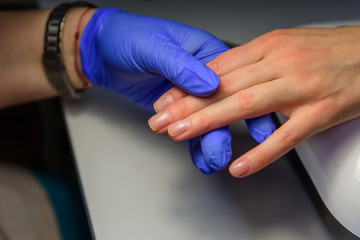Professional manicure in salon, applying the base under the varnish. Hands of master in gloves and fingers of client, glare on nails, close-up.