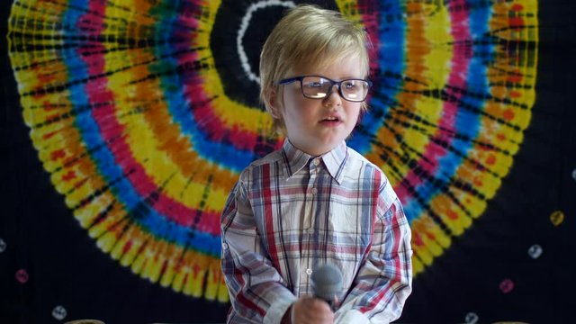 Blond boy with glasses in checkered shirt sings into karaoke microphone. Baby is happy on eve of holiday