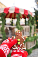 Hand in red mitten holding a smiling gingerbread man and christmas mood in blurred background. Christmas market in old town European small city.