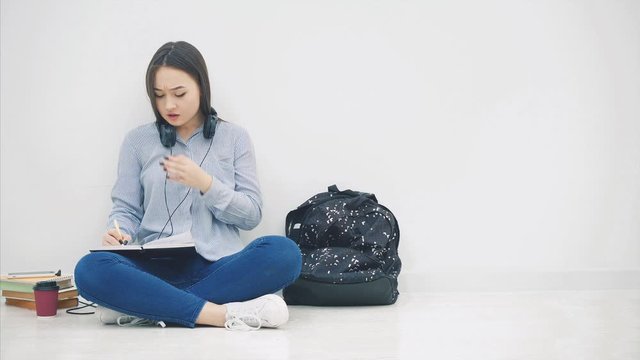 Young asian girl sitting on the floor, is bored of writing, yawning. So, she takes phone and headphones and is listening to music, dancing.