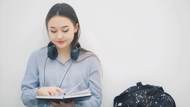 Young asian girl sitting on the floor, is bored of writing, yawning. So, she takes phone and headphones and is listening to music, dancing.