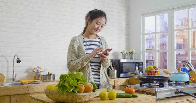 Picture of beautiful asian chinese woman cooking vegetables in cozy wooden kitchen while looking recipe on smartphone. young girl browsing website on tutor cooking class and smiling checking fruits.