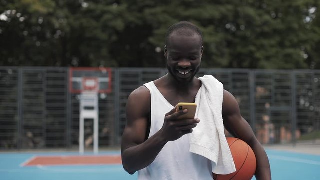 Young Muscly Smiling Afro - American Guy in White Singlet Holding Ball in one hand and Using his Smartphone with other Walking at Street Basketbal Court. Healthy Lifestyle and Sport Concept.