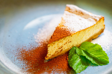 One piece of fresh cheesecake with powdered sugar, cocoa and mint leaf served on a blue plate.