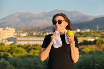 Mature woman in sportswear eating green apple, exercising on nature