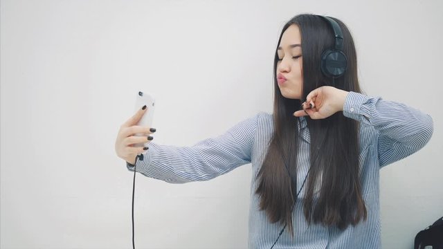 Funny young asian girl standing in headphones, listening to the music, takes selfie, posing, making faces, then messaging with someone.