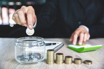 businessman holding coins putting in glass jar,  step of coins stacks on working table, concept saving money for finance accounting.