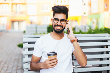 Young freelancer indian man sitting on bench, talking by phone, looking at camera. Handsome man in glasses wearing in white stylish shirt. It worker smiling, posing on background of modern city.