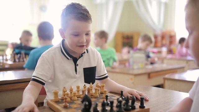 A boy in a white T-shirt is playing chess. Children play educational games in a preschool. Two boy playing chess in light room