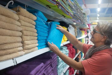 Side view of a smiling senior woman choosing a towel inside a shopping mall. Many colors around her. Consumerism concept. Sales and discounts period