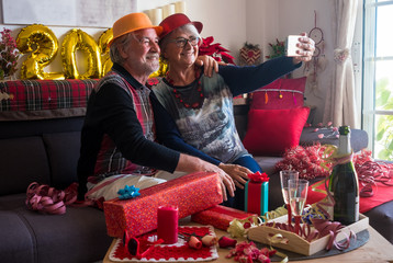 Cheerful couple of senior people with  hat sitting on the sofa and looking at mobile phone for a selfie. Toasting with a sparkling wine. Two people