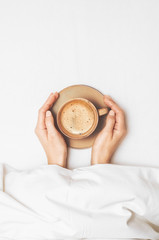 Female hands are holding a cup of morning coffee on the background of a white bedsheet under the covers in bed. Concept wake. Top view. Flat lay