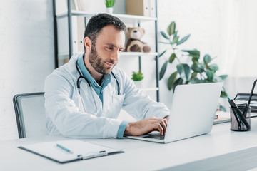 positive doctor sitting at workplace and using laptop - Powered by Adobe