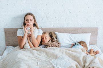 sick woman sneezing in napkin while sitting in bed near diseased children