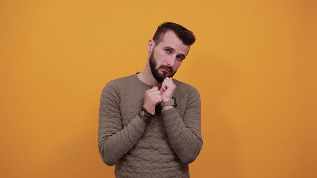 Young cheerful man over isolated orange background looking at camera, keeping hand on chin, pretty wearing casual green clothes