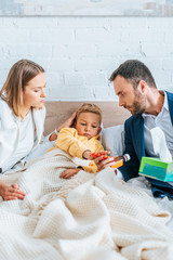 worried father holding cough syrup and mother touching head of diseased son lying in bed