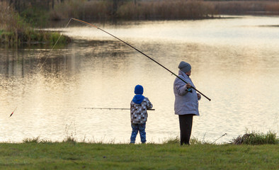 The boy with the grandmother catches fish in the pond at sunset