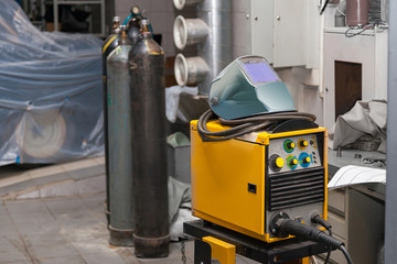 A yellow welding machine with a protective mask on top stands in a metal workshop near carbon...