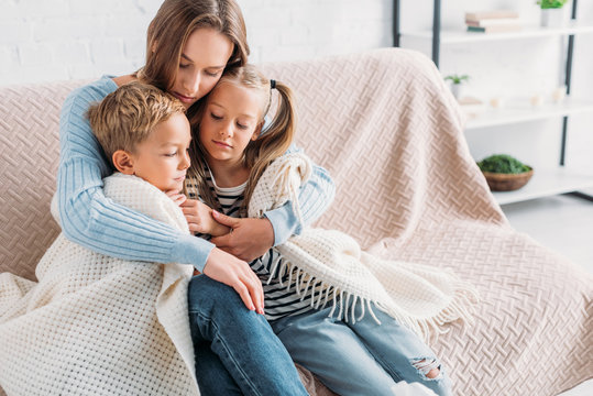 Caring mother wrapping sick children in blanket while sitting on sofa