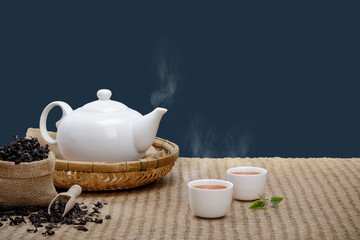 Warm cup of tea with teapot, green tea leaves and dried herbs on the bamboo mat at morning isolate dark blue background with empty space, Organic product from the nature for healthy with traditional