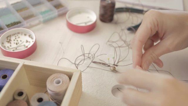 Lifestyle concept, work from home to reinvent your life: close-up detail of woman hands making macrame knotted jewel with some colored thread spools in foreground and bokeh effect filmed in flat color