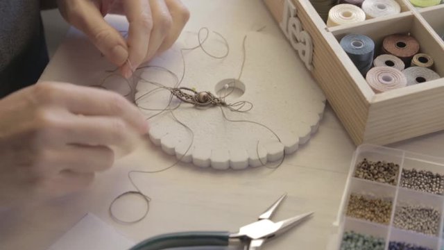 Lifestyle concept, work from home to reinvent your life: close-up detail of woman hands making macrame knotted jewel with some colored thread spools in foreground and bokeh effect filmed in flat color