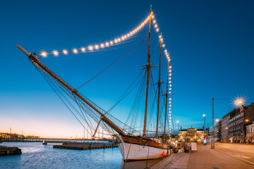 Helsinki, Finland. Old Wooden Sailing Vessel Ship Is Moored To The City Pier, Jetty. Unusual Cafe Restaurant In City Center In Lighting At Evening Or Night Illumination