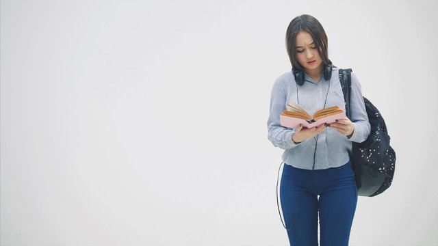 An asian schoolgirl with a backpack appearing on a white background, reading a book, sighing, looking unhappy.