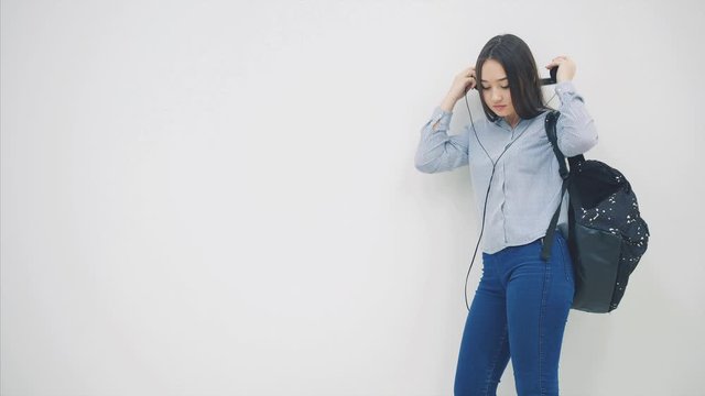 An asian schoolgirl with a backpack appearing on a white background in headphones, taking out and reading notebook.