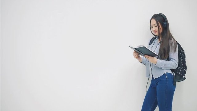 An asian schoolgirl with a backpack appearing on a white background, opening and reading a book, looking happy.