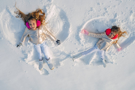 Two Girls On A Snow Angel Shows. Smiling Children Lying On Snow With Copy Space. Funny Kids Making Snow Angel. Top View.