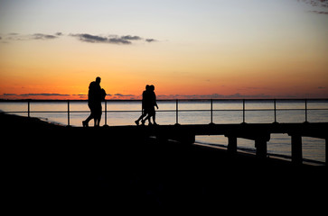 People silhouetted at sunset while enjoying an evening walk along a seaside jetty.
