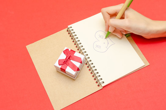 hand of a girl who draws a heart in a notebook, next to a gift box on a red background. concept of Valentine's Day, holiday
