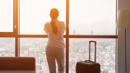 Young brunette woman traveler with suitcase in hotel room with panoramic city view. She is standing...