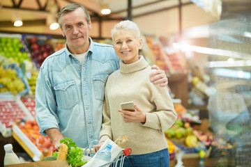 Waist up portrait of modern senior couple pushing cart and smiling at camera while enjoying grocery shopping in supermarket, copy space