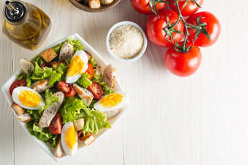 Fresh Caesar salad with delicious chicken breast, ruccola, spinach, cabbage, arugula, egg, parmesan and cherry tomato on wooden background. Oil, salt and pepper. Healthy and diet food concept.