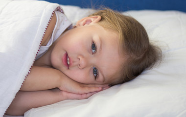 Caucasian child of three age lying in the bed with her hand under the head looking at camera trying to sleep during day