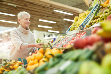 Waist up portrait of smiling adult woman choosing fresh vegetables while grocery shopping at...