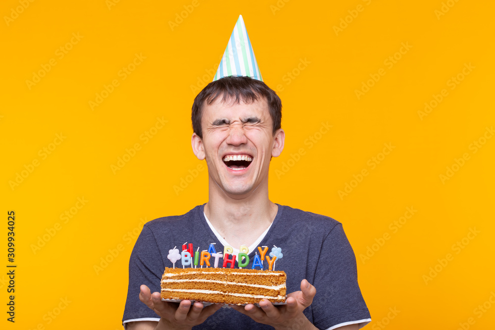 Wall mural Positive funny young asian guy with a cap and a burning candle and a homemade cake in his hands posing on a yellow background. Anniversary and birthday concept.