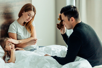 careful father show teddy bear to ill daughter lying on bed while mother checking temperature of...