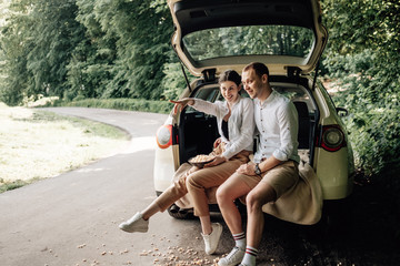 Young Happy Couple Dressed Alike in White T-shirt Sitting in the Car Trunk with Popcorn on the Roadside, Weekend Outside the City, Holidays and Road Trip Concept