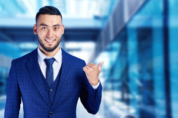 Asian Kazakh businessman in suit smiles friendly and shows finger to the sides on copy space on a blue blurred background