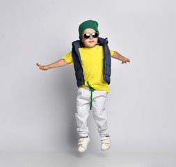 Stylish cheerful little boy 4 years old in sweatpants, a green hat, glasses and a down vest has fun