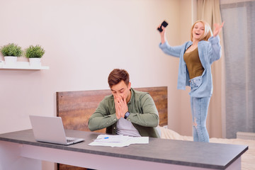 busy man in casual wear sit working on laptop with documents while his wife jumping on bed, happy blond woman indoors