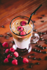 The glass cup of latte coffee with ice, raspberry and coffee grains decoration stands on wooden table 